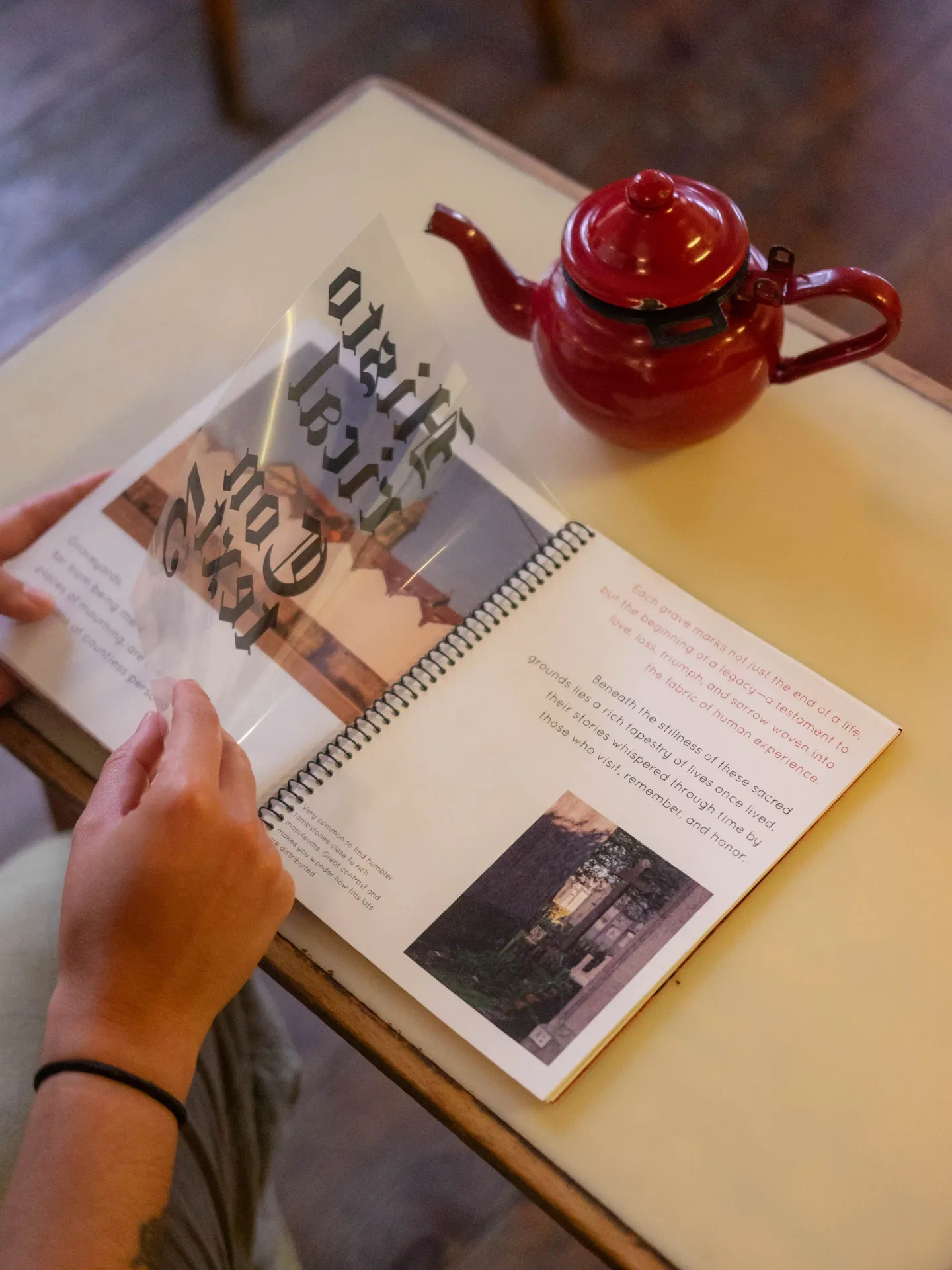 Hands flipping a translucent vellum page of the spiral-bound book, red enamel teapot alongside