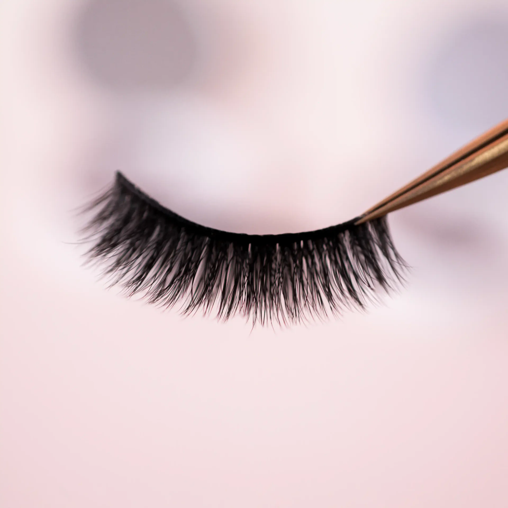 Close-up of a false lash strip held by gold tweezers on a soft pink background