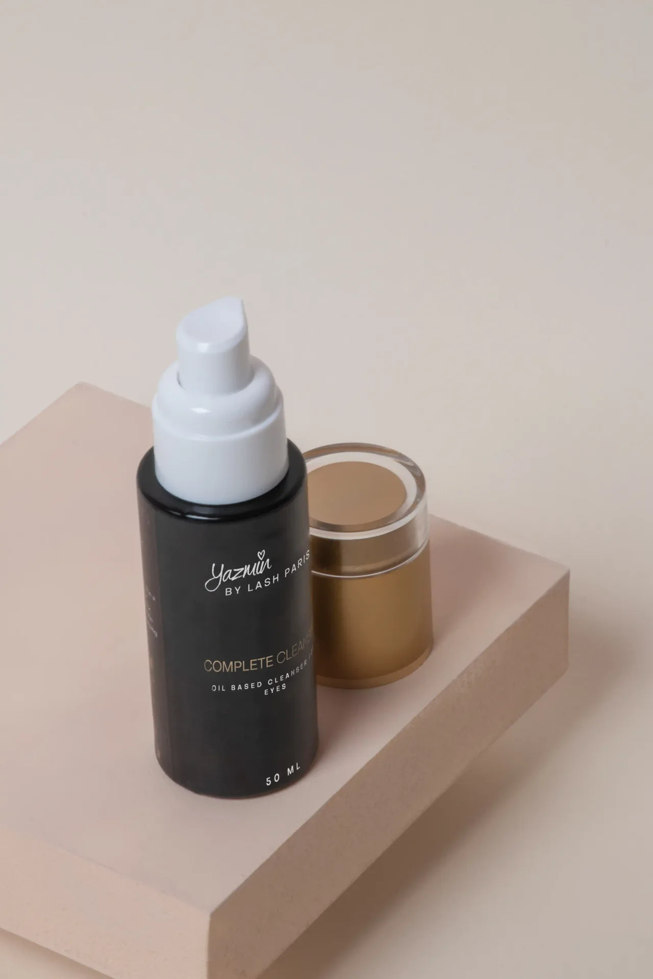 Yazmin by Lash Paris oil-based eye cleanser pump bottle beside a gold jar on a beige plinth