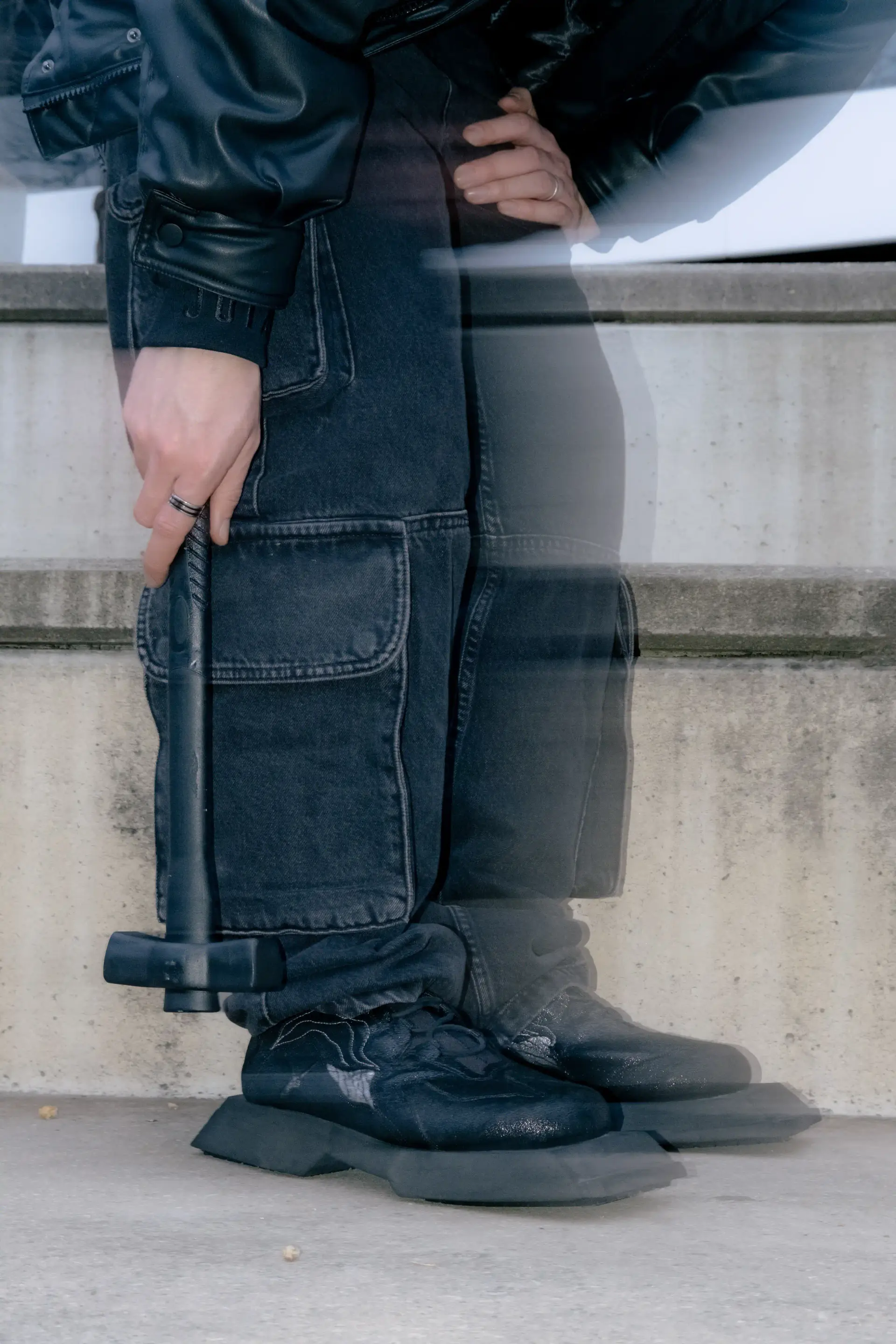 Model in a black leather jacket holding a black mallet beside the sneakers on concrete steps
