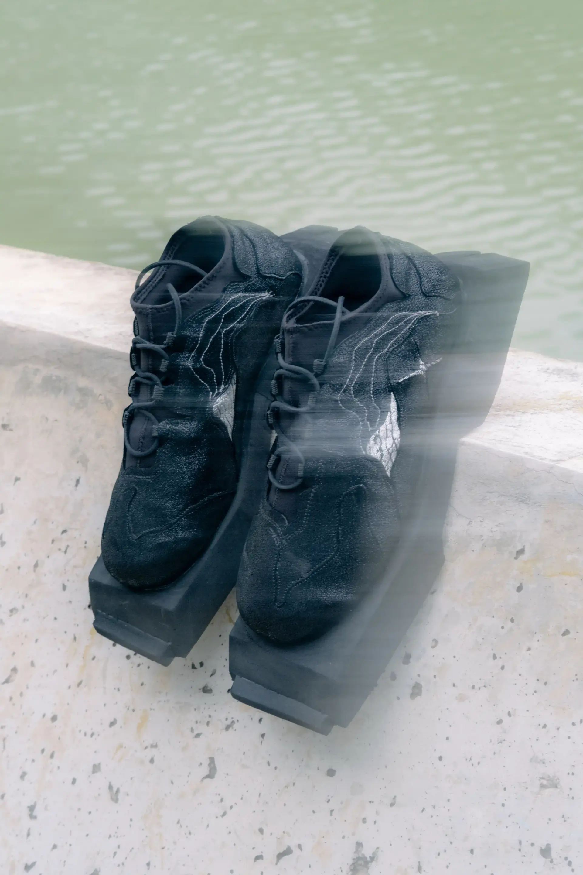 Pair of black Oppressus sneakers with chunky square soles on a concrete ledge above green water