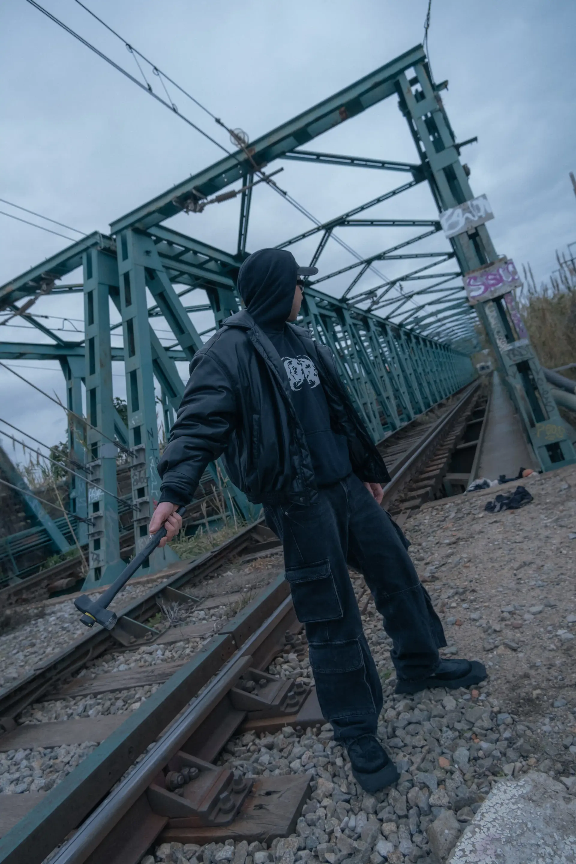 Model in black hood and cap carrying a mallet across a teal steel railway bridge