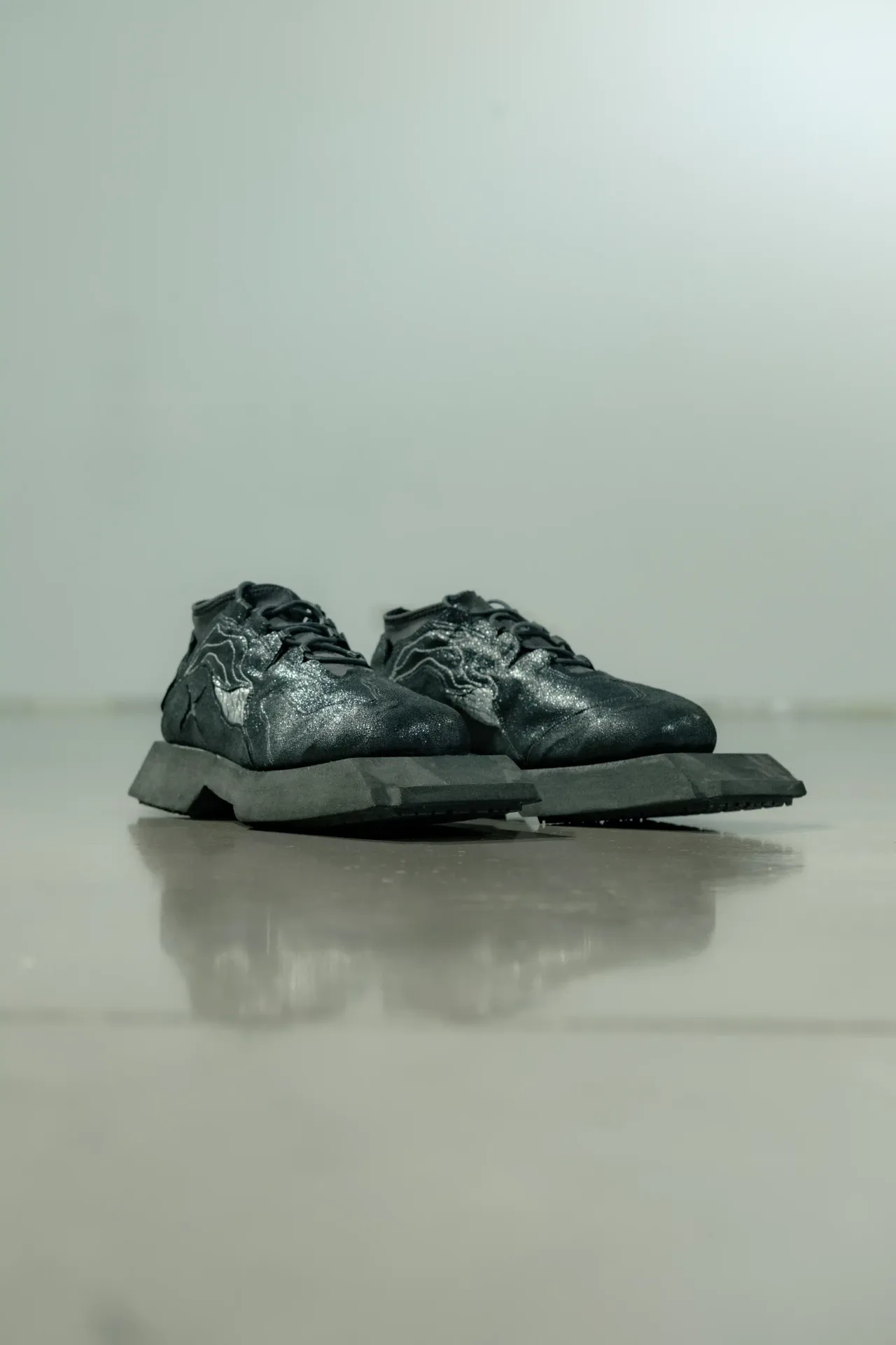 Black Oppressus sneakers with silver stitched detailing on a reflective studio floor
