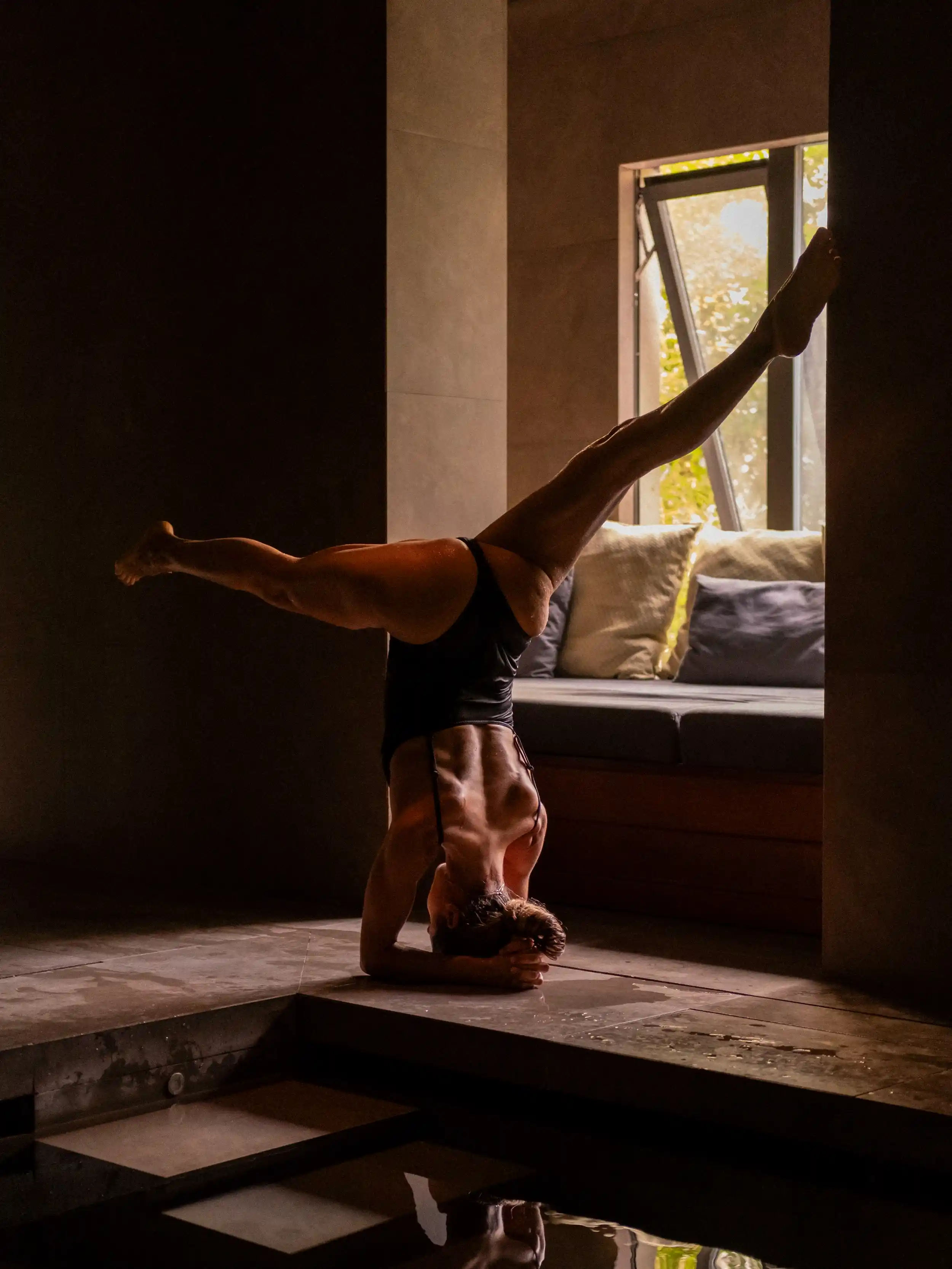 Model holding a headstand yoga pose with legs extended, on the tiled edge of a spa pool